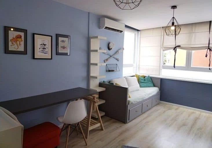 Красивая уютная комната с мягким диваном, яркими подушками и светлой отделкой, окна с жалюзи, стильное освещение и декоративные элементы для комфортного проживания.