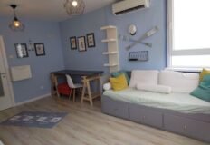 оригинальный интерьер гостиной с уютной мебелью и морской тематикой, светлая палитра и удобный диван с яркими подушками, стильные настенные украшения, большие окна для естественного освещения.