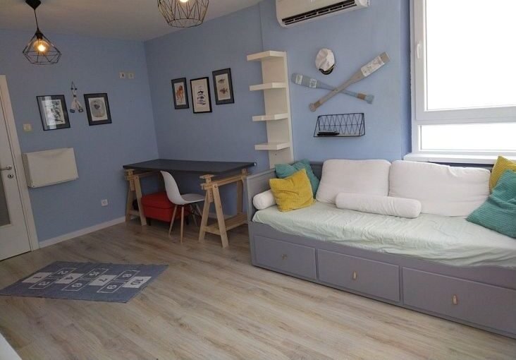 оригинальный интерьер гостиной с уютной мебелью и морской тематикой, светлая палитра и удобный диван с яркими подушками, стильные настенные украшения, большие окна для естественного освещения.