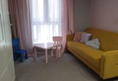 уютная гостиная комната с яркой желтой мягкой мебелью и детскими игрушками, светлый дизайн интерьера, большие окна с легкими шторами, приятная атмосфера для отдыха и развлечений в квартире Eurodomestate.com.